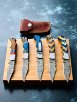 set of 5 Damascus pocket knives with colorful handles on a wooden block, with a leather sheaths one with pocket clip labeled 'ZB Knives' in the background.